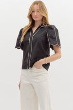 Milan Piped Blouse, a sophisticated black button-down top with statement puff sleeves and contrasting white piping along the collar, front, and sleeves. The structured yet flowy silhouette adds a modern touch to a classic design.